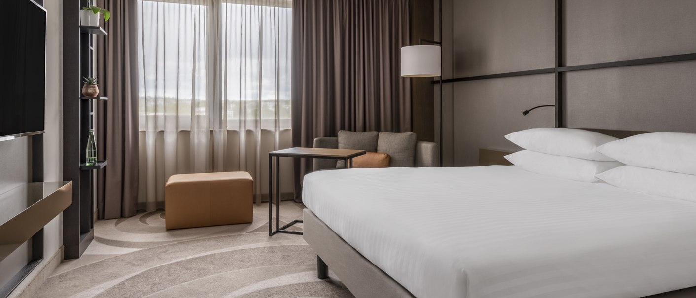 Modernes Doppelzimmer mit großem Bett, Sofa, Tisch und Fenster mit Vorhängen. Dezente Farben und stilvolle Einrichtung., © Stuttgart Marriott Hotel Sindelfingen