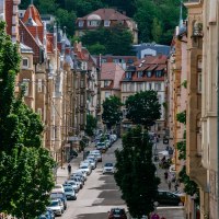 Eine belebte Stadtstra&szlig;e mit geparkten Autos, alten Geb&auml;uden und B&auml;umen. Im Hintergrund sind H&uuml;gel und weitere H&auml;user zu sehen., &copy; Thomas Niederm&uuml;ller