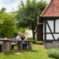 Drei Personen sitzen an einem Holztisch neben einem kleinen Fachwerkhaus im Gr&uuml;nen. Es ist ein sonniger Tag., &copy; Corinna Jacobs