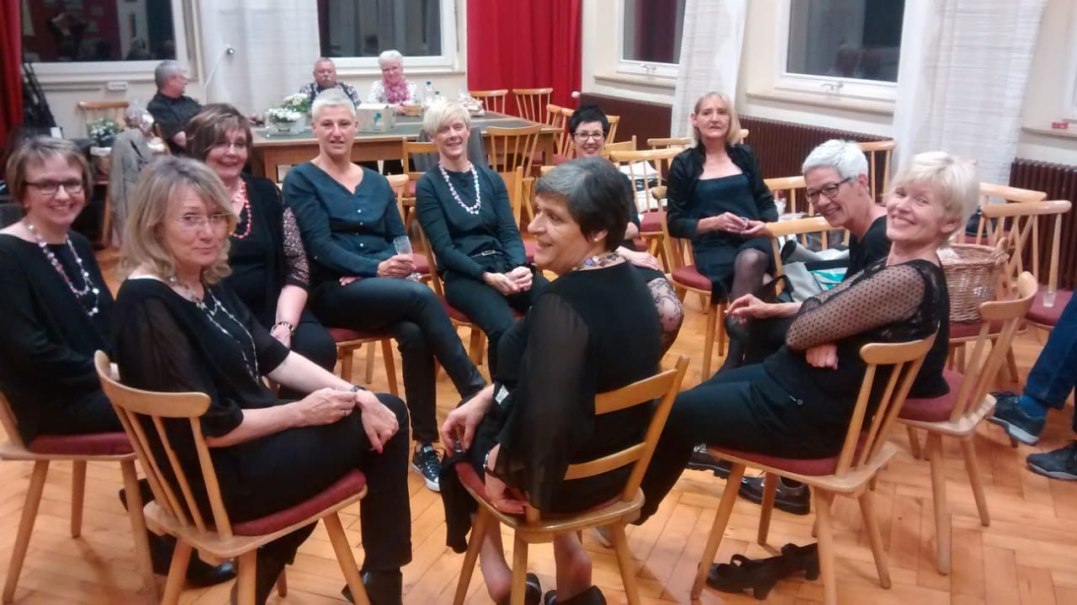 Frauen in schwarzer Kleidung sitzen l&auml;chelnd in einem Kreis auf St&uuml;hlen in einem Raum mit Holzboden und roten Vorh&auml;ngen., &copy; TonArt