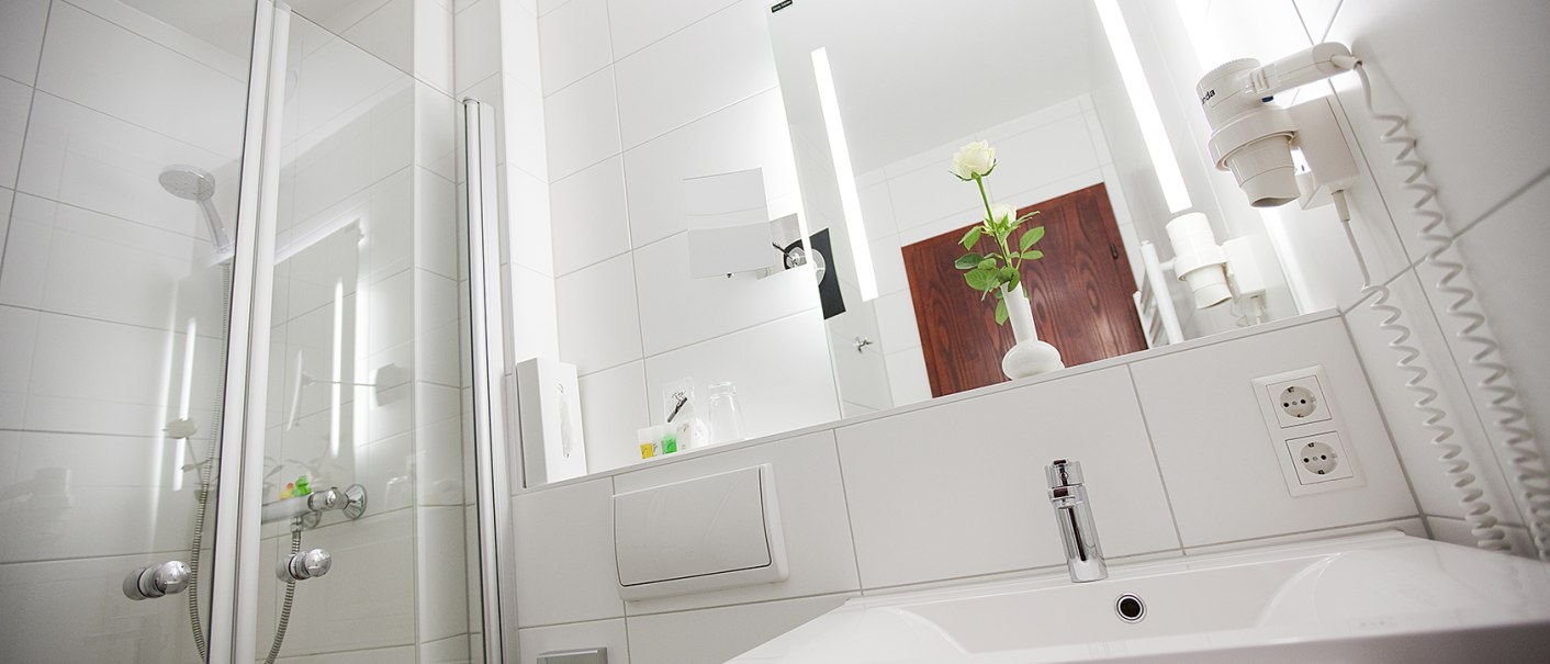 Modernes Badezimmer mit weißer Fliesenwand, Dusche, Waschbecken, Spiegel und einer weißen Blume in einer Vase., © Hotel Krone Modernes Badezimmer mit weißer Fliesenwand, Dusche, Waschbecken, Spiegel und einer weißen Blume in einer Vase., © Hotel Krone