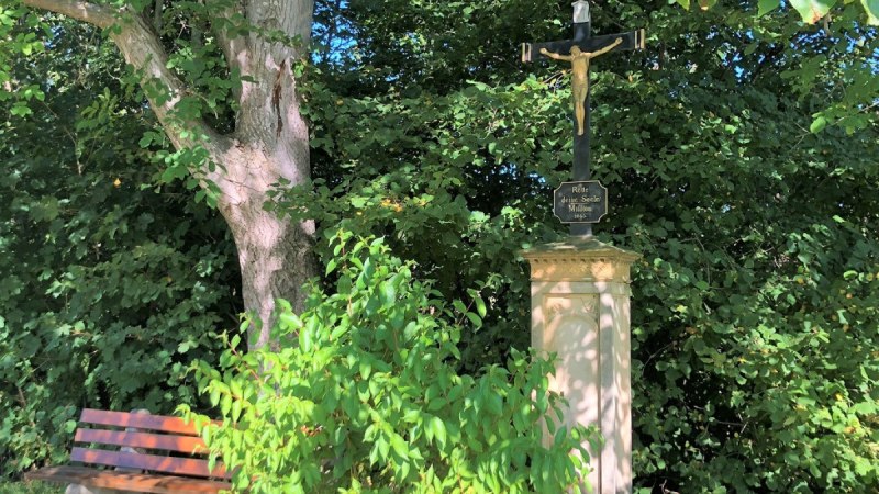 Ein steinernes Kreuz mit Jesusfigur steht neben einer Holzbank unter einem Baum, umgeben von dichtem Grün., © Touristik und Marketing GmbH Ein steinernes Kreuz mit Jesusfigur steht neben einer Holzbank unter einem Baum, umgeben von dichtem Grün., © Touristik und Marketing GmbH