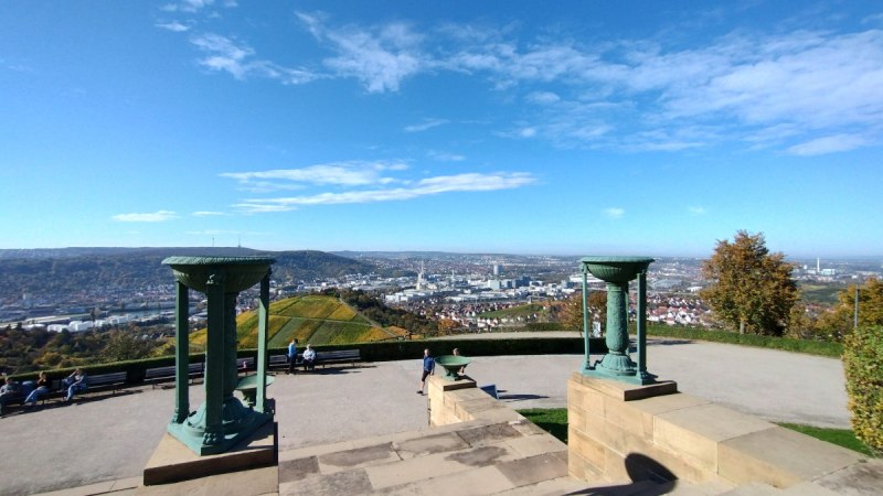 Panoramablick vom Rotenberg auf Stuttgart, umgeben von Weinbergen. Zwei dekorative Säulen im Vordergrund, blauer Himmel und vereinzelte Wolken.