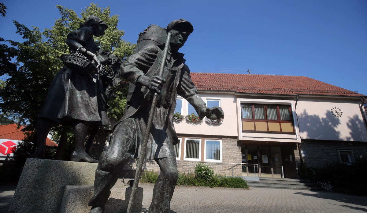 Bronzestatue eines Mannes und einer Frau vor dem Rathaus Neuhausen. Das Gebäude hat ein rotes Dach und helle Wände, umgeben von Bäumen.