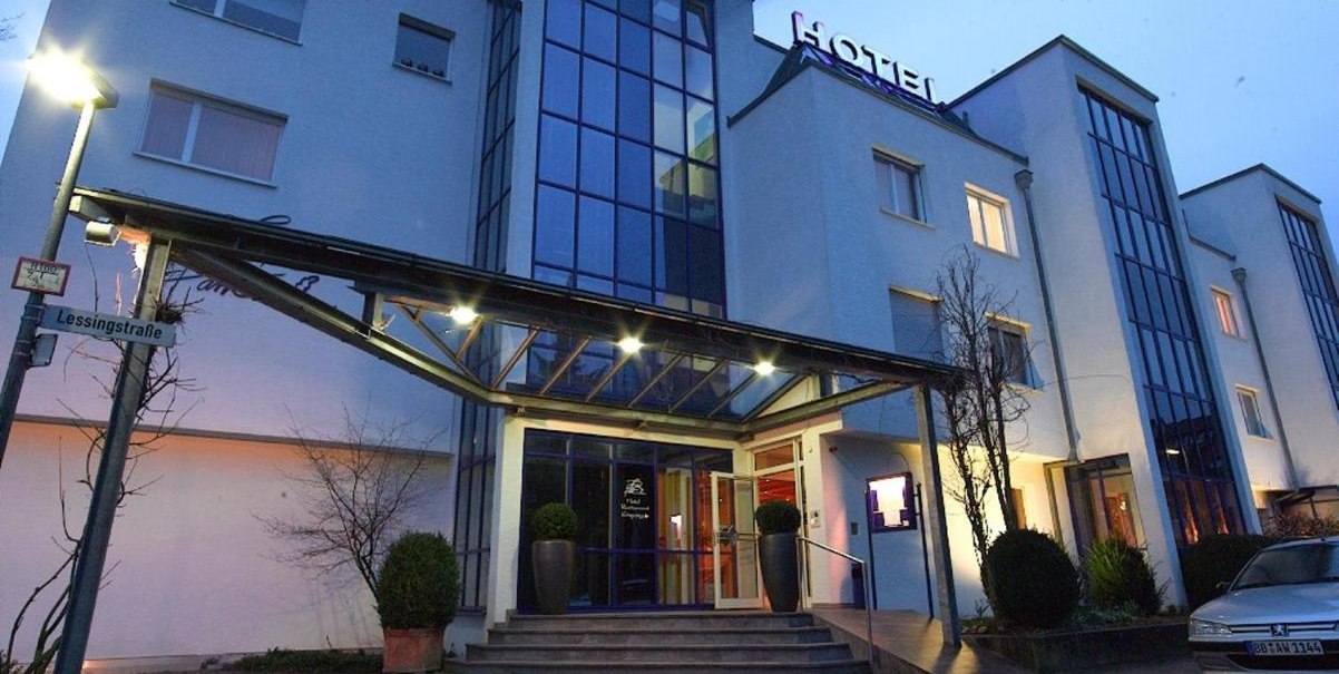 Modernes Hotel mit Glasfassade und beleuchtetem Eingang bei D&auml;mmerung. Ein Stra&szlig;enschild zeigt 'Lessingstra&szlig;e'. Ein Auto parkt vor dem Geb&auml;ude., &copy; Hotel am Park