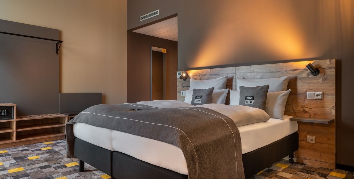 Modernes Hotelzimmer mit Doppelbett, Holzkopfteil, grauen Kissen und gemustertem Teppich in Grau und Gelb., © Novum Hospitality Modernes Hotelzimmer mit Doppelbett, Holzkopfteil, grauen Kissen und gemustertem Teppich in Grau und Gelb., © Novum Hospitality