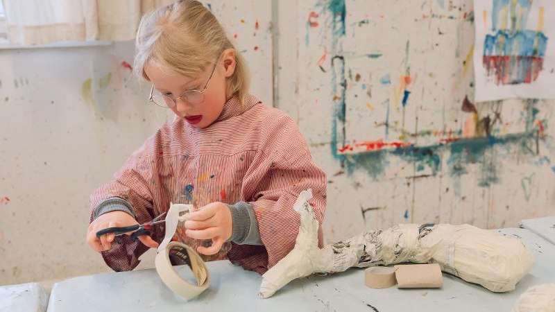 Ein Kind in einem gestreiften Kittel bastelt konzentriert mit Klebeband und Schere. Der Hintergrund zeigt eine kreative, farbenfrohe Umgebung., &copy; Kunstschule Labyrinth