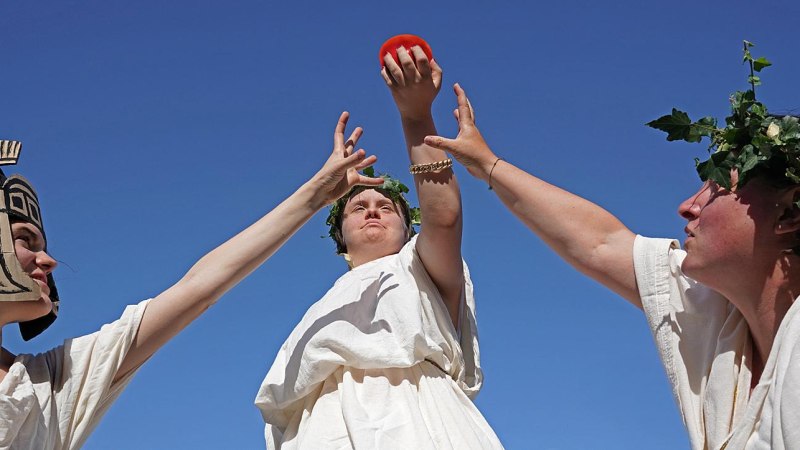 Drei Personen in antiken Kostümen strecken die Hände nach einem roten Objekt aus. Eine trägt einen Helm, die anderen Blätterkränze. Blauer Himmel im Hintergrund., © Albrecht Fendrich Drei Personen in antiken Kostümen strecken die Hände nach einem roten Objekt aus. Eine trägt einen Helm, die anderen Blätterkränze. Blauer Himmel im Hintergrund., © Albrecht Fendrich