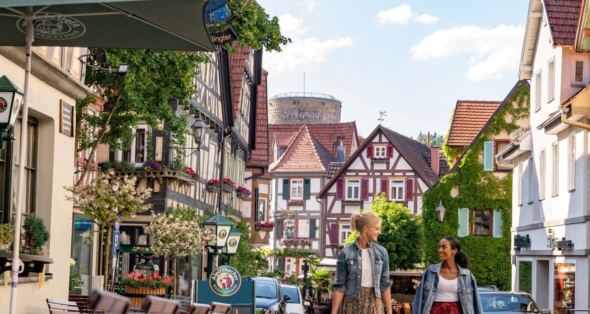 Zwei Frauen gehen eine gepflasterte Straße entlang, gesäumt von Fachwerkhäusern und einem Schild des Ratsstüble in Besigheim., © Stuttgart Marketing GmbH Zwei Frauen gehen eine gepflasterte Straße entlang, gesäumt von Fachwerkhäusern und einem Schild des Ratsstüble in Besigheim., © Stuttgart Marketing GmbH