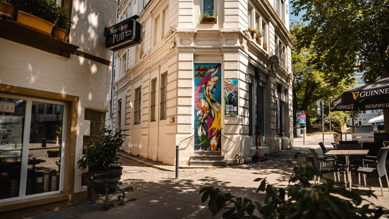 Eine Straßenecke mit einem Pub-Schild, bunter Wandkunst und Café-Tischen unter einem Sonnenschirm., © SMG Stuttgart Marketing GmbH - Sarah Schmid Eine Straßenecke mit einem Pub-Schild, bunter Wandkunst und Café-Tischen unter einem Sonnenschirm., © SMG Stuttgart Marketing GmbH - Sarah Schmid
