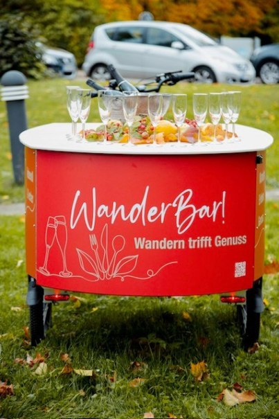 Ein mobiler Tisch mit Gl&auml;sern und Snacks, beschriftet mit 'WanderBar! Wandern trifft Genuss', steht auf einer Wiese.