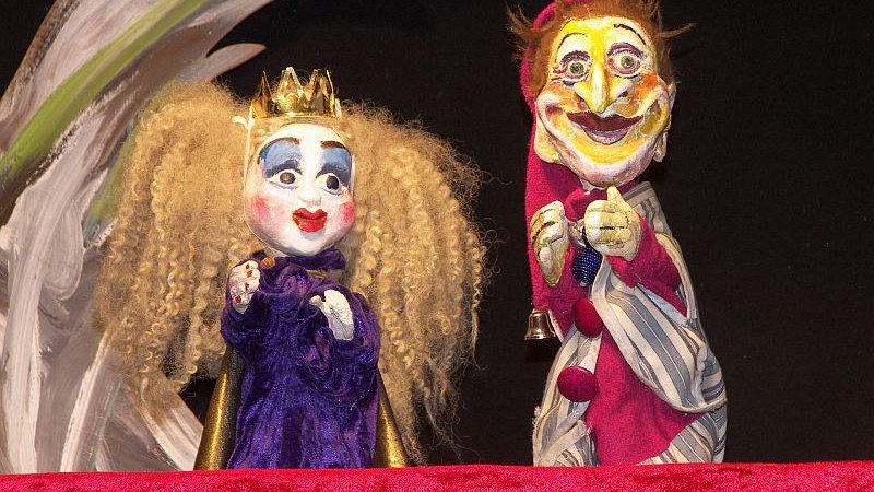Zwei Handpuppen, eine Königin mit blonden Locken und Krone, und ein Kasperl mit roter Mütze, stehen vor einem bemalten Hintergrund auf einer Bühne., © Backnang Zwei Handpuppen, eine Königin mit blonden Locken und Krone, und ein Kasperl mit roter Mütze, stehen vor einem bemalten Hintergrund auf einer Bühne., © Backnang