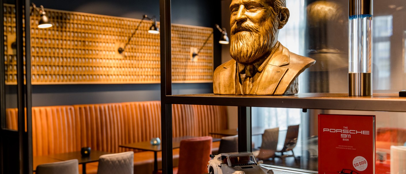 Büste eines Mannes mit Bart, ein Modellauto und ein Porsche-Buch in einem stilvoll eingerichteten Raum mit Ledersitzbänken., © TOMAS
