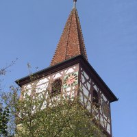Ein Kirchturm mit Fachwerk und Uhr ragt über Bäume in den blauen Himmel. Der Turm hat ein spitzes Dach mit Wetterhahn., © Natur.Nah. Schönbuch & Heckengäu