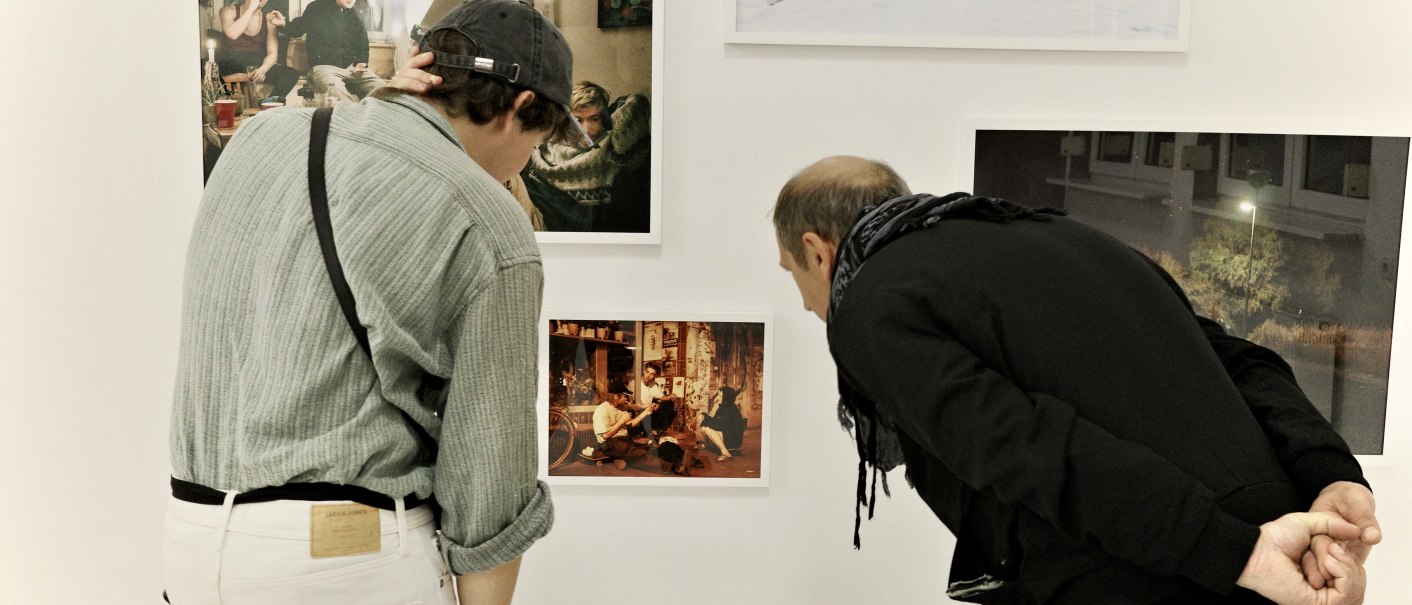 Zwei Personen betrachten aufmerksam Fotos an einer weißen Galeriewand. Die Bilder zeigen verschiedene Szenen, darunter Menschen und eine Schneelandschaft., © Palermo Galerie Zwei Personen betrachten aufmerksam Fotos an einer weißen Galeriewand. Die Bilder zeigen verschiedene Szenen, darunter Menschen und eine Schneelandschaft., © Palermo Galerie