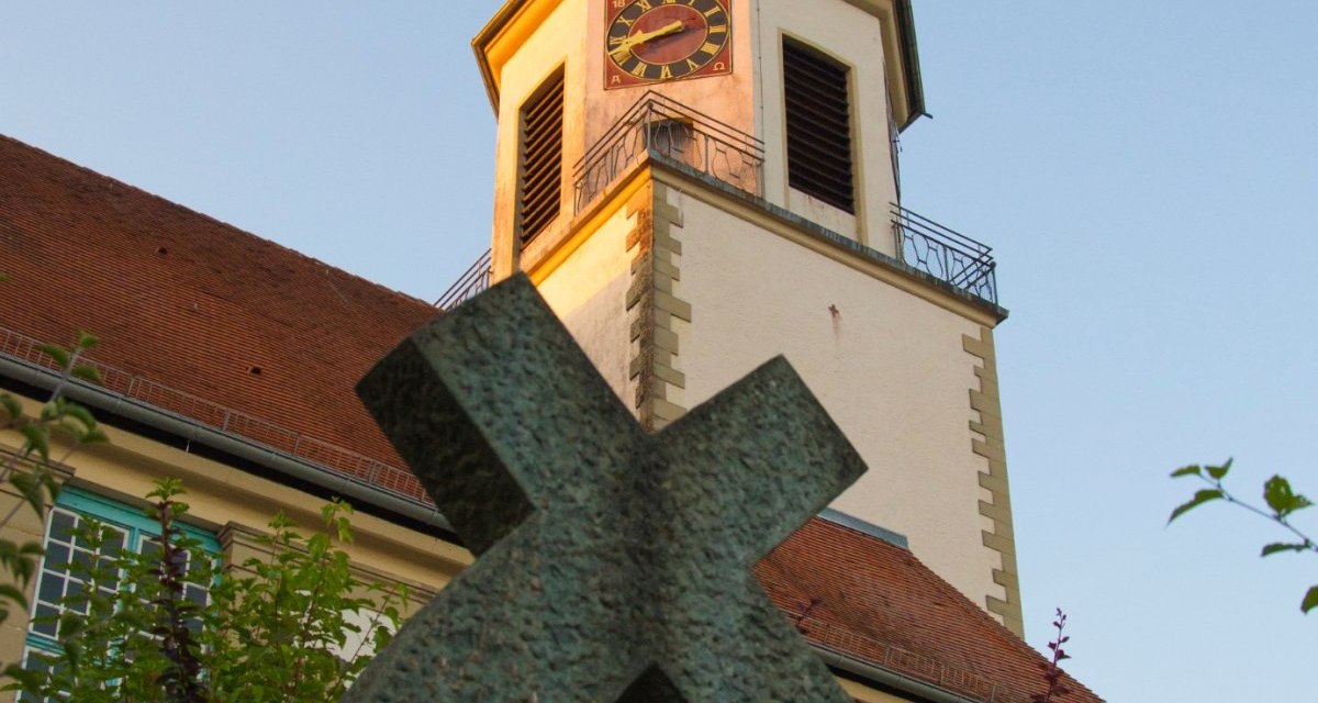 Ein Kirchturm mit Uhr und Wetterhahn ragt in den blauen Himmel. Im Vordergrund steht ein schiefes Kreuz, umgeben von grünen Pflanzen., © Unbekannt Ein Kirchturm mit Uhr und Wetterhahn ragt in den blauen Himmel. Im Vordergrund steht ein schiefes Kreuz, umgeben von grünen Pflanzen., © Unbekannt