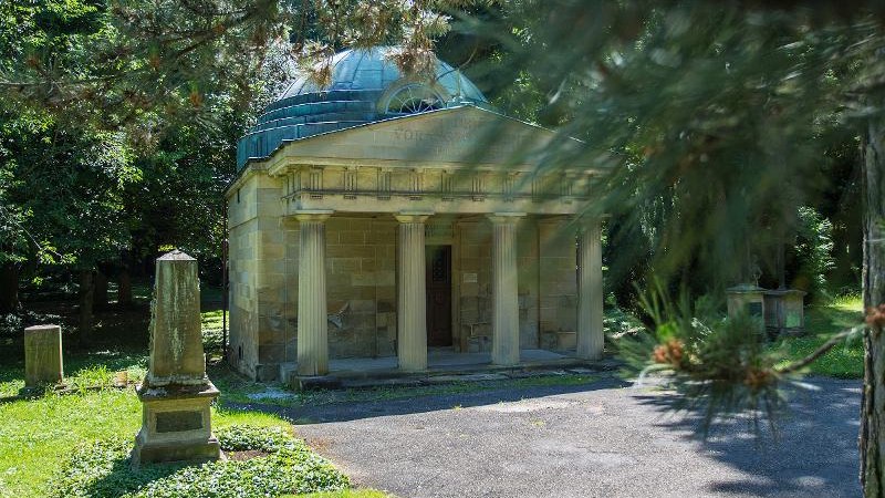 Ein altes Mausoleum mit Säulen und Kuppel auf einem Friedhof, umgeben von Bäumen und Grabsteinen. Die Sonne scheint durch die Äste., © © T&E, Benjamin Stollenberg Ein altes Mausoleum mit Säulen und Kuppel auf einem Friedhof, umgeben von Bäumen und Grabsteinen. Die Sonne scheint durch die Äste., © © T&E, Benjamin Stollenberg