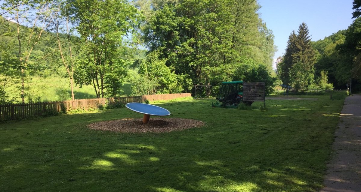 Ein ruhiger Spielplatz mit einem runden Spielgerät, umgeben von grüner Natur und Bäumen. Ein Traktor steht im Hintergrund., © www.pro-cycl.de Ein ruhiger Spielplatz mit einem runden Spielgerät, umgeben von grüner Natur und Bäumen. Ein Traktor steht im Hintergrund., © www.pro-cycl.de
