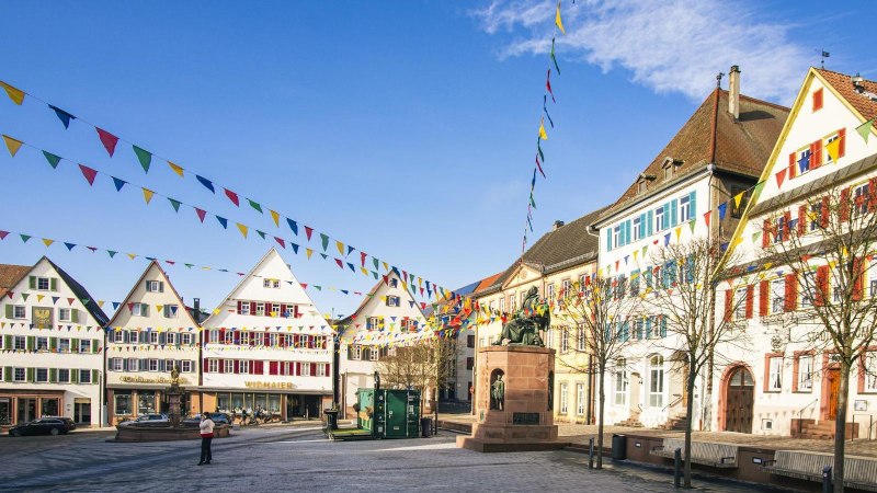 Der Marktplatz von Weil der Stadt zeigt historische Gebäude und bunte Wimpel unter blauem Himmel. Eine Statue steht zentral auf dem Platz., © SMG, Sarah Schmid Der Marktplatz von Weil der Stadt zeigt historische Gebäude und bunte Wimpel unter blauem Himmel. Eine Statue steht zentral auf dem Platz., © SMG, Sarah Schmid