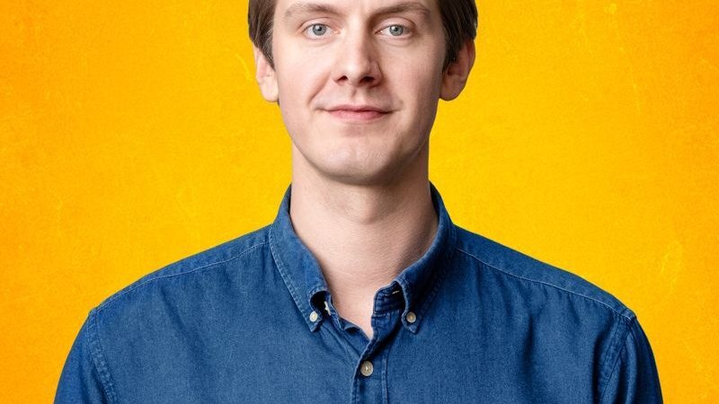 Ein Mann in blauem Hemd und beiger Hose sitzt lächelnd auf dem Boden vor einem grauen Hintergrund., © Stadt Leinfelden-Echterdingen, Kulturamt