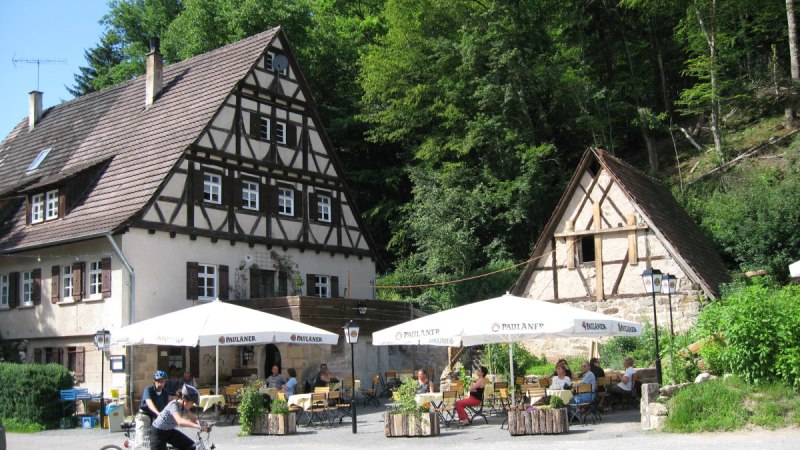 Ein Fachwerkhaus mit Biergarten, umgeben von Bäumen. Menschen sitzen an Tischen, während zwei Radfahrer im Vordergrund stehen., © Natur.Nah. Schönbuch & Heckengäu