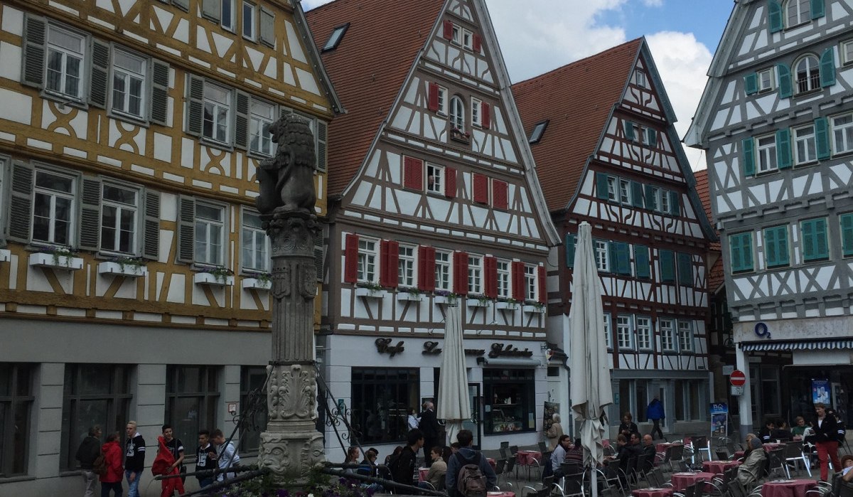 Fachwerkhäuser am Marktplatz in Herrenberg, Deutschland. Ein Brunnen steht im Vordergrund, umgeben von Menschen und Cafétischen. Der Himmel ist blau mit Wolken., © www.pro-cycl.de Fachwerkhäuser am Marktplatz in Herrenberg, Deutschland. Ein Brunnen steht im Vordergrund, umgeben von Menschen und Cafétischen. Der Himmel ist blau mit Wolken., © www.pro-cycl.de