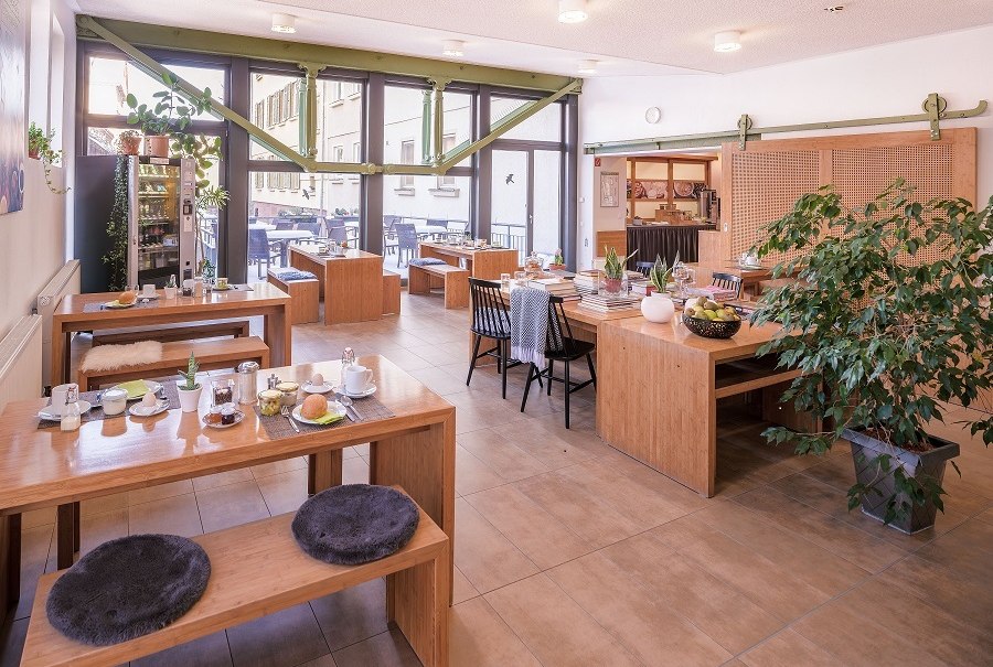 Heller Fr&uuml;hst&uuml;cksraum mit Holztischen, Pflanzen und gro&szlig;en Fenstern. Gem&uuml;tliche Atmosph&auml;re mit gedeckten Tischen und Sitzkissen., &copy; ECOINN Hotel am Campus