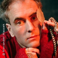 Person in rotem Pullover hält ein Blatt und eine Kette vor dem Gesicht, schaut direkt in die Kamera. Hintergrund ist dunkel., © links im Bild Person in rotem Pullover hält ein Blatt und eine Kette vor dem Gesicht, schaut direkt in die Kamera. Hintergrund ist dunkel., © links im Bild