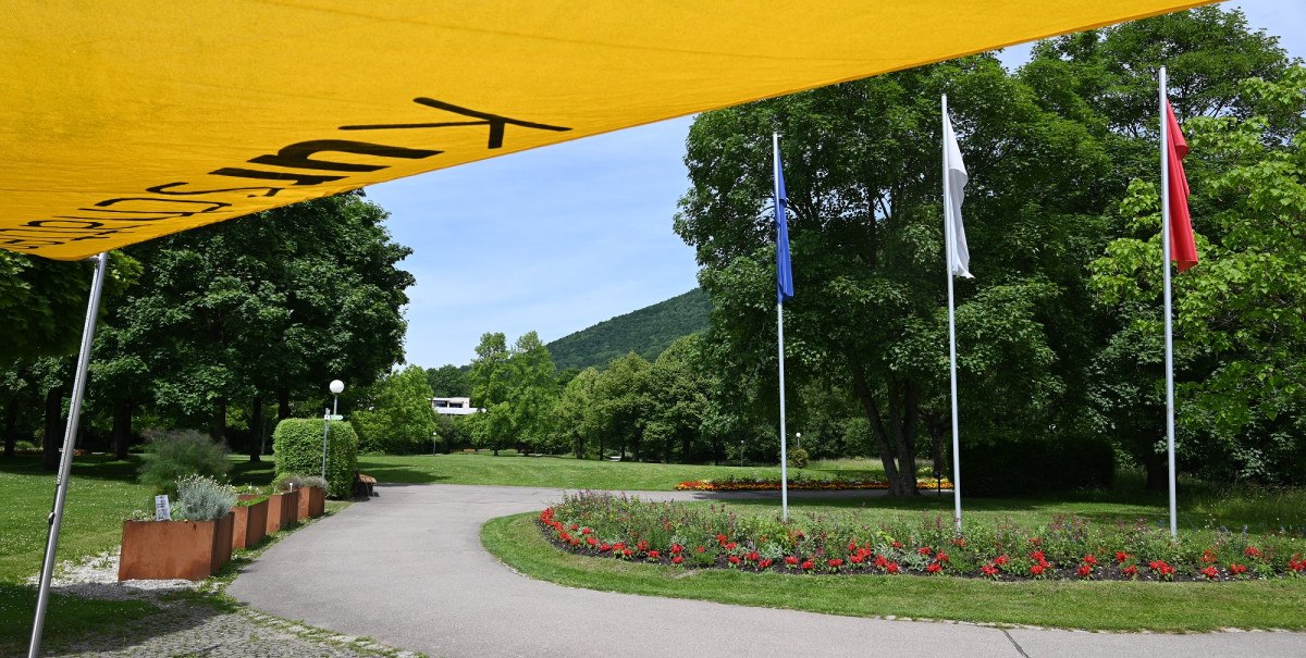 Kurpark Bad Urach mit gelbem Sonnensegel, drei Flaggen und gepflegten Blumenbeeten entlang eines Weges.