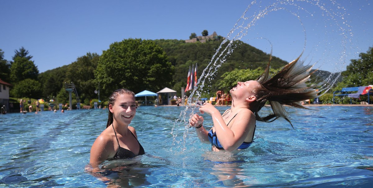 Zwei M&auml;dchen im Freibad, eines wirft Wasser mit den Haaren in die Luft. Im Hintergrund sind B&auml;ume und ein H&uuml;gel zu sehen.