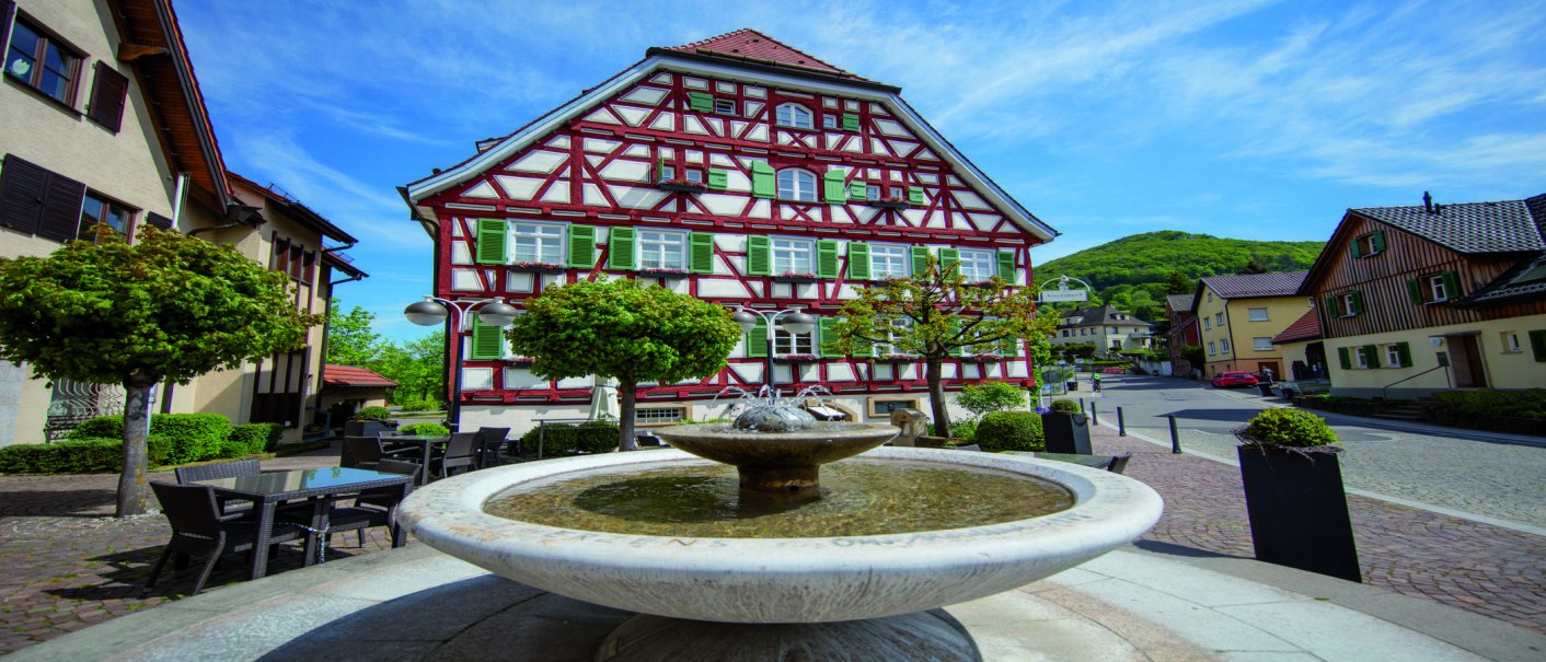 Fachwerkhaus in Bad Überkingen mit grün-weißen Fensterläden, umgeben von Bäumen und einem Brunnen im Vordergrund. Blauer Himmel im Hintergrund., © © Stuttgart-Marketing GmbH