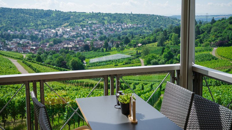 Terrasse mit Tisch und Stühlen, Blick auf grüne Weinberge und ein Dorf im Hintergrund. Sonniger Tag, klare Sicht auf die Landschaft., © Rotenberger Weingärtle, Frederik Garlin