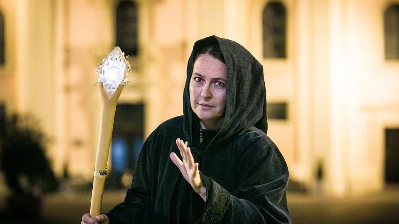 Eine Person in einem schwarzen Umhang hält einen leuchtenden Stab und steht vor einem unscharfen Gebäude bei Nacht., © © T&E, Benjamin Stollenberg Eine Person in einem schwarzen Umhang hält einen leuchtenden Stab und steht vor einem unscharfen Gebäude bei Nacht., © © T&E, Benjamin Stollenberg