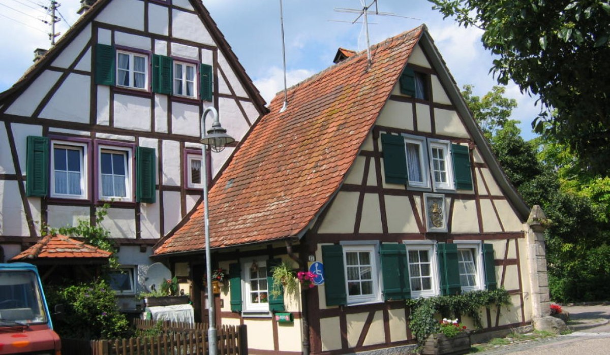 Zwei Fachwerkhäuser mit roten Ziegeldächern und grünen Fensterläden, umgeben von Pflanzen und einem geparkten blauen Fahrzeug., © Land der 1000 Hügel - Kraichgau-Stromberg Zwei Fachwerkhäuser mit roten Ziegeldächern und grünen Fensterläden, umgeben von Pflanzen und einem geparkten blauen Fahrzeug., © Land der 1000 Hügel - Kraichgau-Stromberg