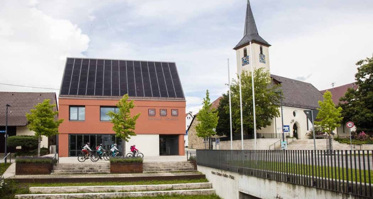 Ein modernes Gebäude mit Solarpanelen und eine Kirche in Jesingen. Vor dem Gebäude fahren Kinder auf Fahrrädern. Im Vordergrund sind Treppenstufen., © Schwäbische Alb Tourismusverband e.V. Ein modernes Gebäude mit Solarpanelen und eine Kirche in Jesingen. Vor dem Gebäude fahren Kinder auf Fahrrädern. Im Vordergrund sind Treppenstufen., © Schwäbische Alb Tourismusverband e.V.