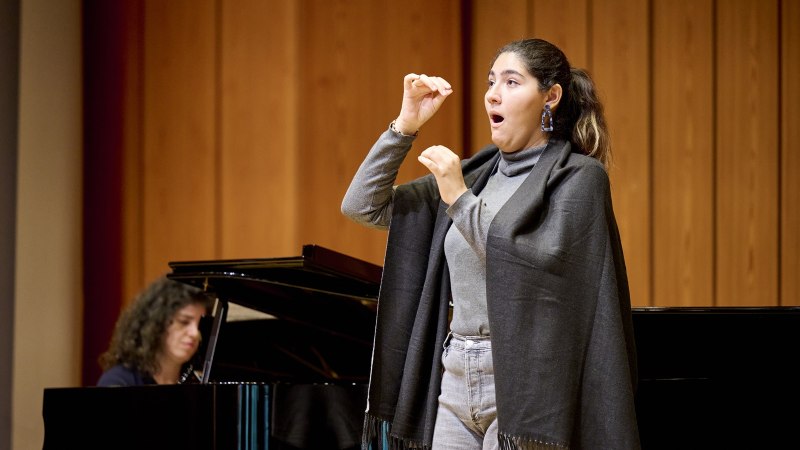 Eine Frau singt leidenschaftlich auf einer B&uuml;hne, w&auml;hrend eine Pianistin im Hintergrund spielt. Der Raum hat eine h&ouml;lzerne Wandverkleidung., &copy; www.peter-oppenlaender.de