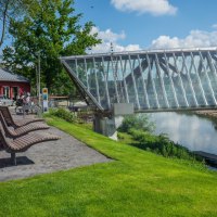 Moderne Glasbrücke über Fluss in Remseck am Neckar, umgeben von grüner Landschaft und einem roten Gebäude. Sitzbänke und Radfahrer im Vordergrund., © Remstal Tourismus e.V.