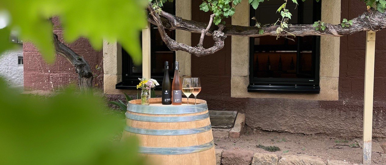 Ein Weinfass dient als Tisch mit zwei Weinflaschen und Gläsern darauf, umgeben von Weinreben vor einem rustikalen Gebäude., © Weingut Schwarz Ein Weinfass dient als Tisch mit zwei Weinflaschen und Gläsern darauf, umgeben von Weinreben vor einem rustikalen Gebäude., © Weingut Schwarz