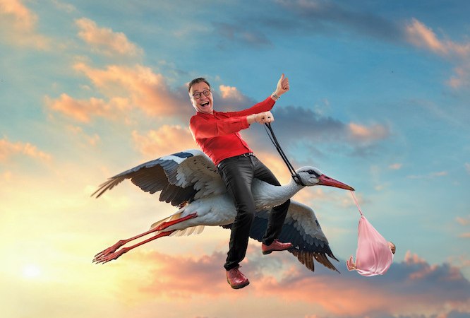 Eine Person in rotem Oberteil reitet l&auml;chelnd auf einem Storch, der ein B&uuml;ndel tr&auml;gt, vor einem farbenfrohen Sonnenuntergangshimmel., &copy; Vaddi Concerts GmbH