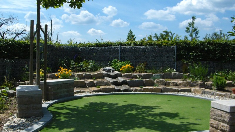 Minigolfplatz mit künstlichem Grün, umgeben von Pflanzen und Steinmauern. Ein Baum spendet Schatten, der Himmel ist blau mit weißen Wolken., © Anja Beyerle & Ralf Bähr GbR