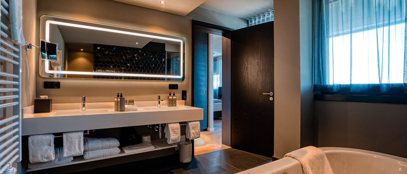 Modernes Badezimmer mit Doppelwaschbecken, beleuchtetem Spiegel und Badewanne. Durch die Tür sieht man ein Schlafzimmer. Elegantes Design mit dunklen Fliesen., © CLOUD N°7 APARTMENTS GmbH