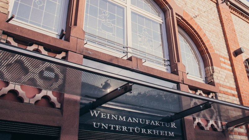 Fassade der Weinmanufaktur Untertürkheim mit Glasvordach, Schriftzug und dekorativen Fenstern., © Weinmanufaktur Stuttgart