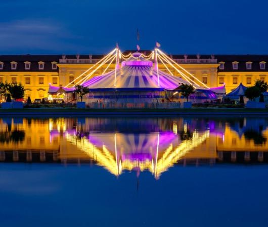 Ein beleuchtetes Zirkuszelt von Circus Roncalli im Bl&uuml;henden Barock, reflektiert in einem Wasserbecken bei Nacht. Die Lichter strahlen in Gelb und Lila., &copy; &copy; Oliver Kelkar