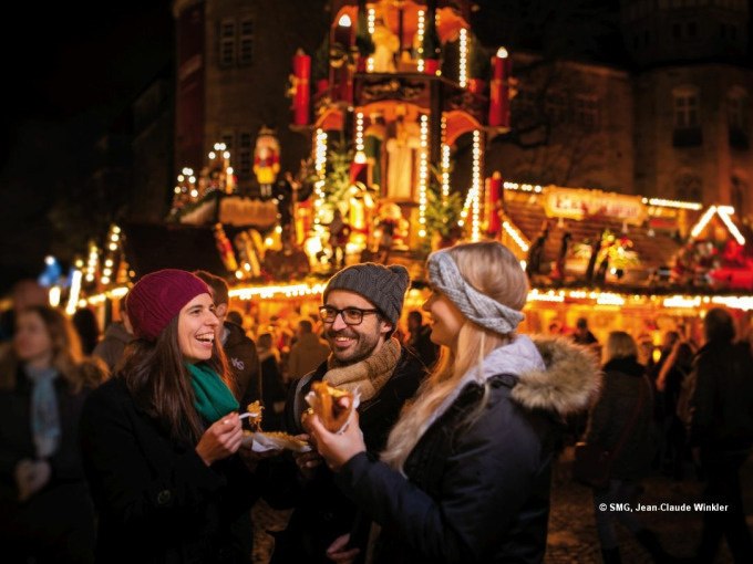 Drei Personen lachen und essen auf einem festlich beleuchteten Weihnachtsmarkt in Stuttgart. Im Hintergrund sind dekorierte Stände zu sehen., © Stuttgart Marketing GmbH Drei Personen lachen und essen auf einem festlich beleuchteten Weihnachtsmarkt in Stuttgart. Im Hintergrund sind dekorierte Stände zu sehen., © Stuttgart Marketing GmbH