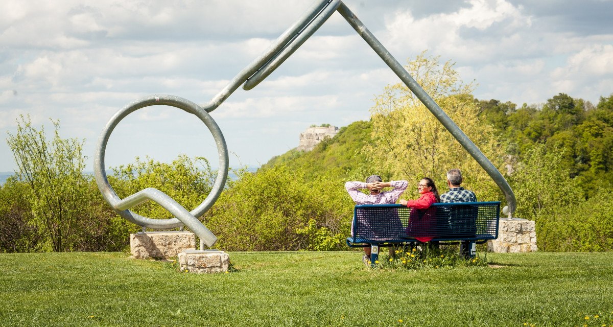 Drei Personen sitzen auf einer Bank vor einer großen Brillenskulptur in einer grünen Landschaft mit Hügeln und Wolken am Himmel., © hochgehberge Drei Personen sitzen auf einer Bank vor einer großen Brillenskulptur in einer grünen Landschaft mit Hügeln und Wolken am Himmel., © hochgehberge