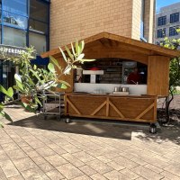 Ein Holzstand vor einem modernen Geb&auml;ude mit Glasfassade. Im Vordergrund sind Pflanzen zu sehen, die den Stand teilweise verdecken., &copy; TOMAS