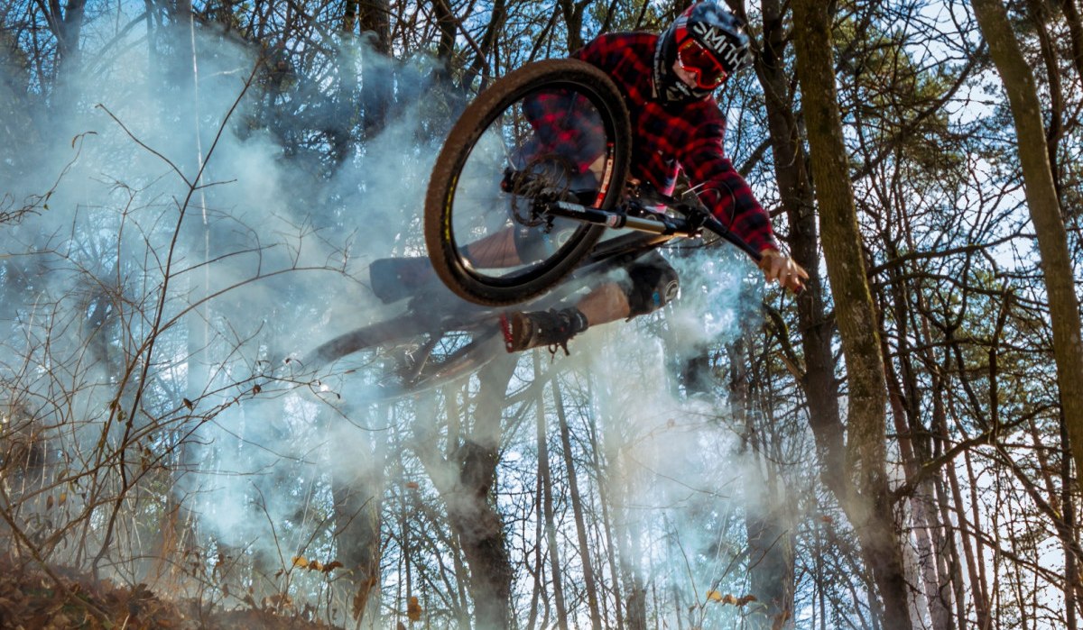 Ein Mountainbiker in rotem Karohemd springt durch Rauch im Wald. Die Szene ist dynamisch und zeigt herbstliche Bäume im Hintergrund., © Natur.Nah. Schönbuch & Heckengäu