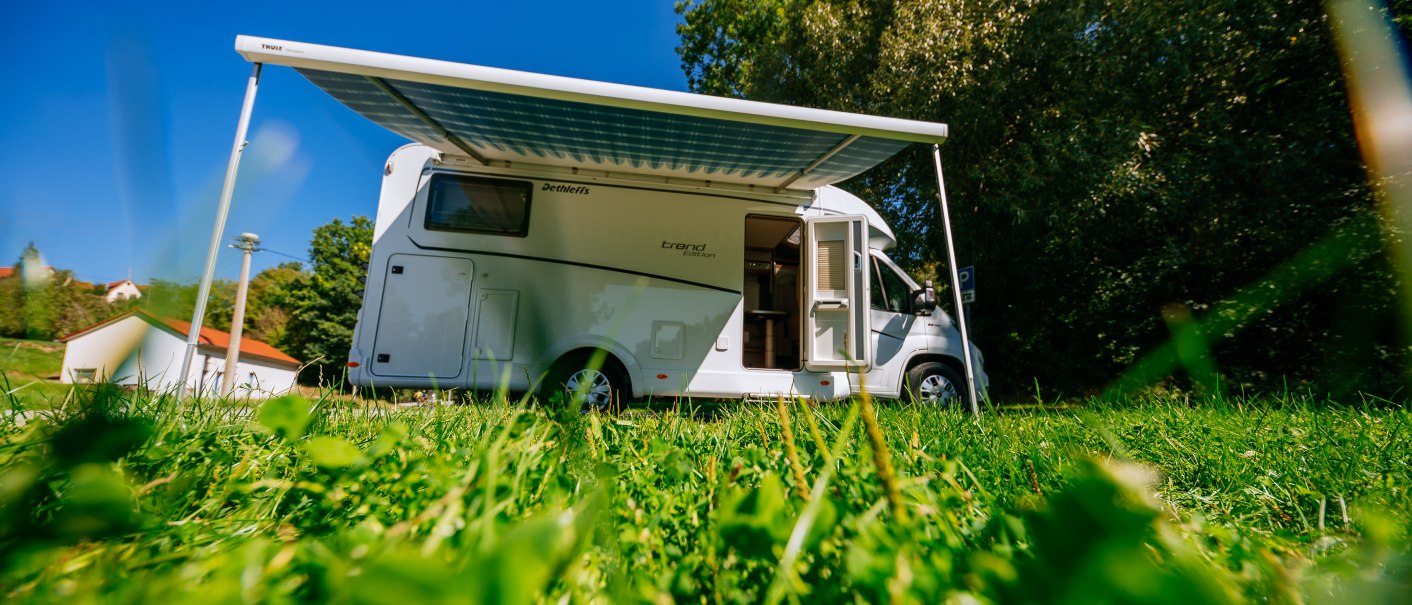 Ein Wohnmobil mit ausgefahrener Markise steht auf einer gr&uuml;nen Wiese unter blauem Himmel. Im Hintergrund sind B&auml;ume und ein Geb&auml;ude zu sehen., &copy; SMG, Thomas Niederm&uuml;ller