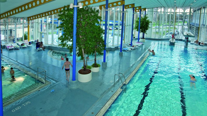 Ein Hallenbad mit mehreren Schwimmbecken, Liegestühlen und großen Fenstern. Menschen schwimmen und entspannen. Pflanzen dekorieren den Raum., © Stuttgart-Marketing GmbH