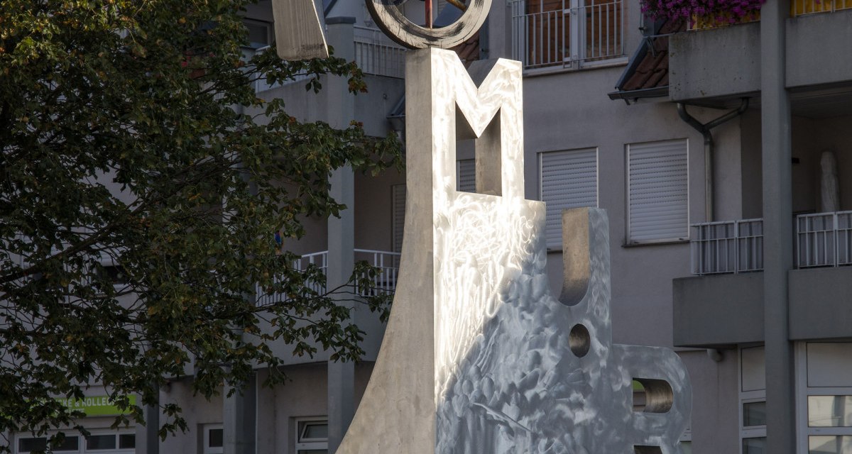 Der Katz- und Mausbrunnen in Bottwartal zeigt eine moderne Skulptur mit Buchstaben und geometrischen Formen vor einem Gebäude mit roten Dachziegeln., © Stuttgart Marketing
